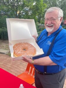 Mark Buckley is Pictured holding up a donut box with one donut inside. The donut is approximately one foot in diameter and glazed.