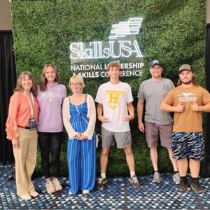 Four students and 2 advisors are shown posing in front of a fake grass wall backdrop with a neon sign that reads