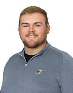 A photo of new Hutch Rec sports coordinator, Taylor Martin. Taylor is a male with short blonde hair, a beard and mustache, and he is wearing a gray pullover with a Kansas Jayhawks logo.