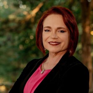 Stacy Rogers is shown in front of a grenn nature backdrop. She has short, vibrant red hair, and is wearing a pink blouse with a black blazer.